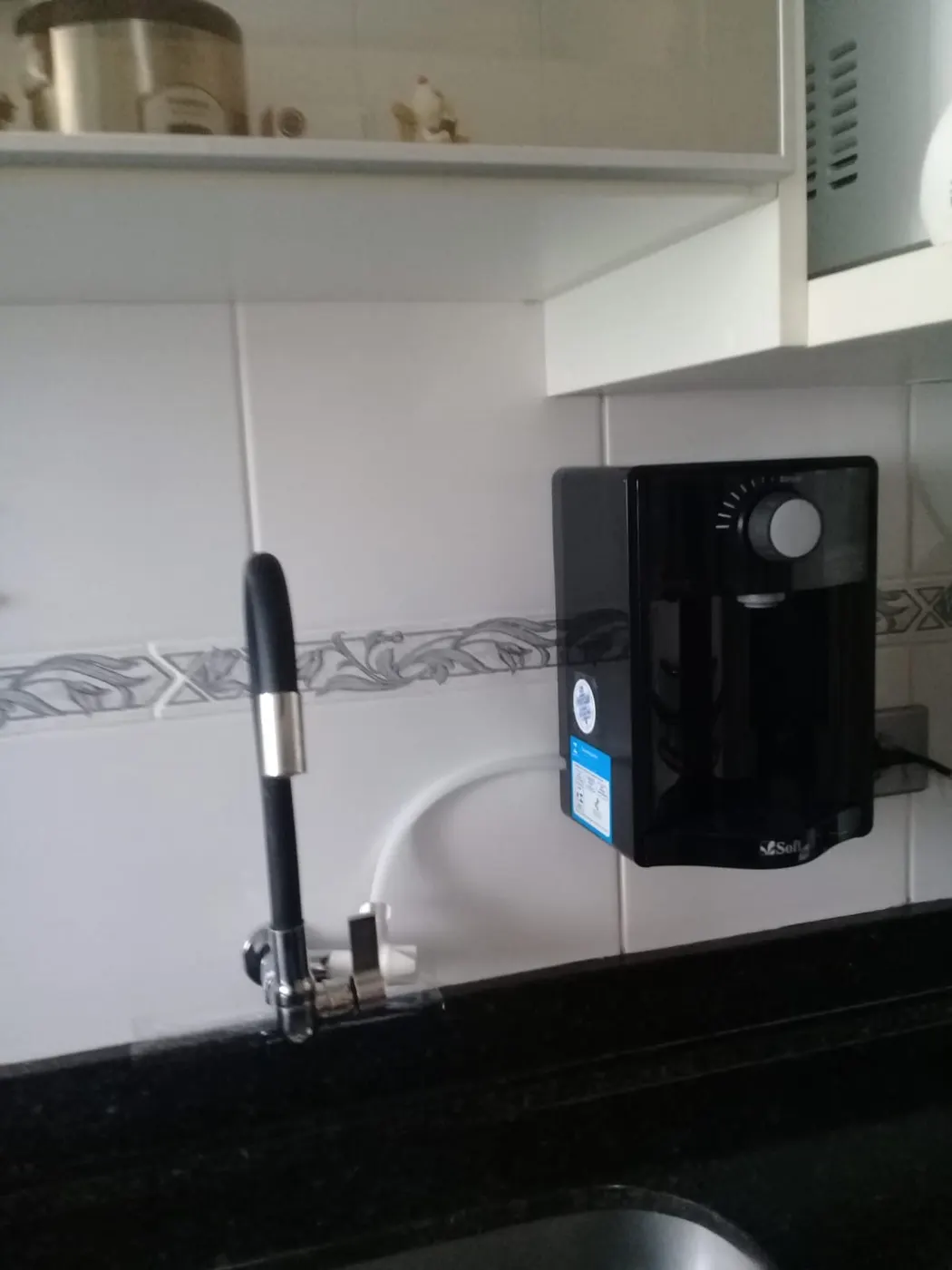 Purificador de água Soft preto instalado em parede de cozinha ao lado de torneira