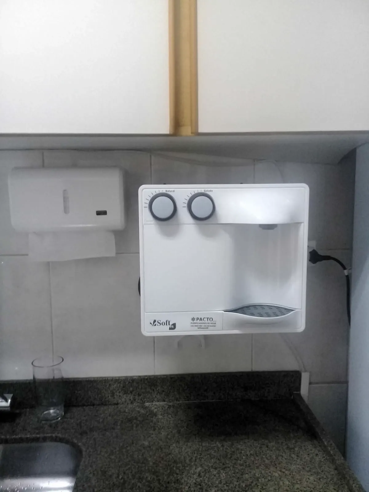 Purificador de água branco instalado em parede de copa ao lado de um porta-papel toalha