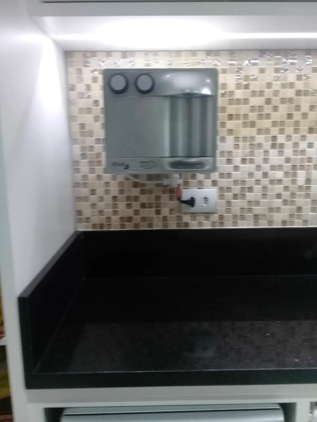 Purificador de água prata instalado em parede de pastilhas acima de bancada de granito preto