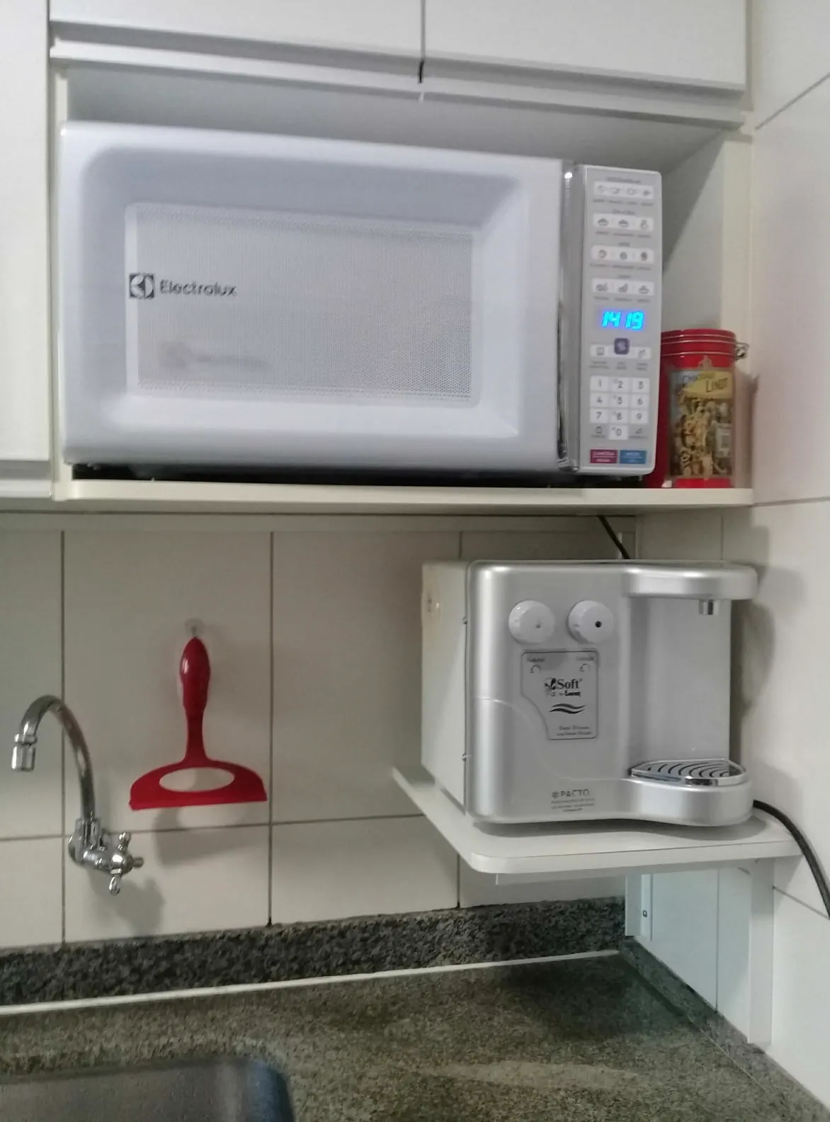 Purificador de água prata apoiado em prateleira branca abaixo de um micro-ondas